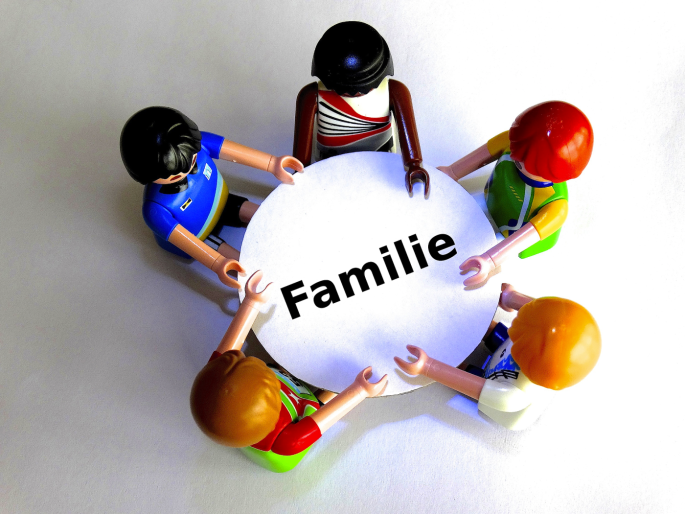 Auf dem Bild sieht man Legofiguren, die an einem Tisch sitzen und sich die Hände geben. Auf der Tischplatte steht das Wort "Familie".