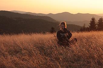 Auf dem Bild sieht man eine junge Frau die in einem Feld steht. Im Hintergrund sind Bäume und Berge. Das Bild ist etwas dunkel und vom Farbton her braun gehalten.