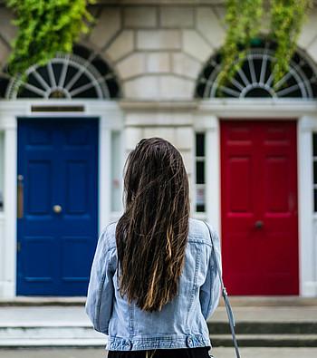 Auf dem Bild sieht man eine brünette Frau mit langen Haaren, die vor zwei Türen steht. Eine ist blau und die andere rot.