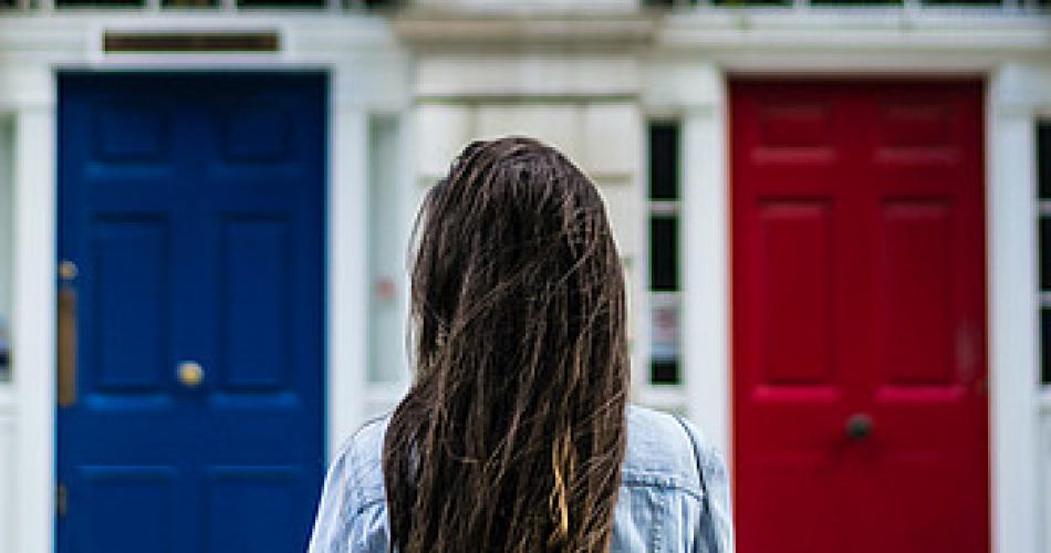 Auf dem Bild sieht man eine brünette Frau mit langen Haaren, die vor zwei Türen steht. Eine ist blau und die andere rot.